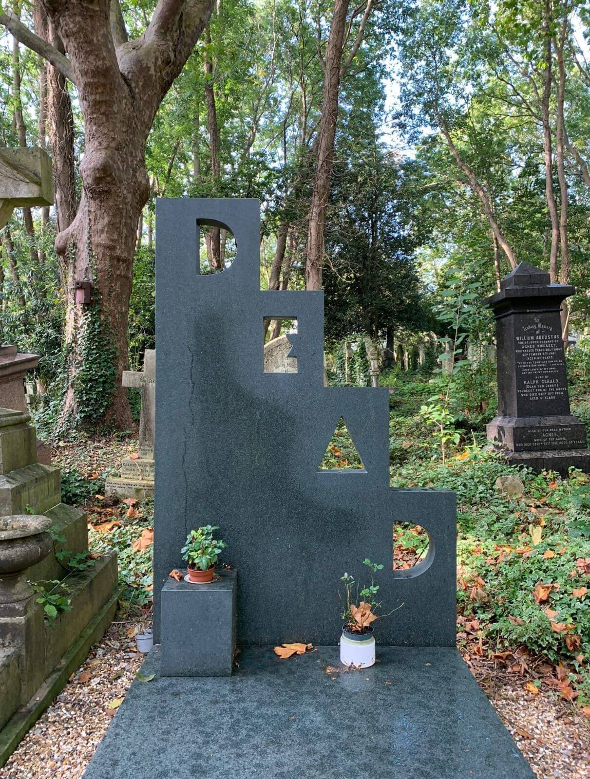 Grey stepped gravestone in a forest, each step containing a letter that form the word DEAD. At the bottom of the grave there are flowers.